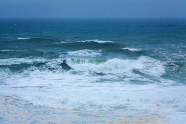 Yuvarlanan dalgalar ve beyaz köpüklerle dolu engin, güçlü bir okyanus yumuşak, sisli bir mavi gökyüzünün altında uzanır ve dinamik bir deniz manzarası yaratır.