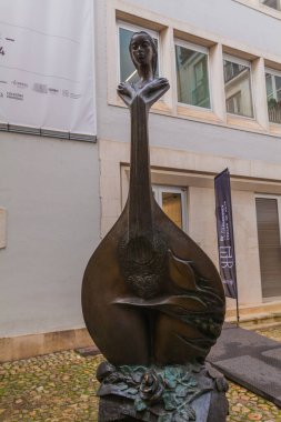 Lavtayla birleşen bronz bir kadın heykeli kaldırımlı bir avluda modern bir binaya karşı duruyor.