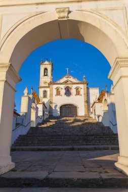 Beyazlatılmış bir cephesi ve öne çıkan bir çan kulesi olan büyük bir kilise, taş bir kemer ve girişe çıkan bir merdivenle çevrili.