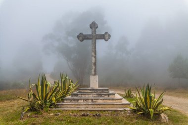 Bir taş haç, puslu bir arazide dimdik durur. Etrafı agave bitkileriyle çevrili ve toprak bir patika.