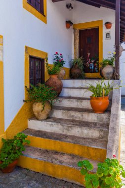 Beyaz yüzlü, sarı süslemeli ve terracotta kiremitli geleneksel bir Portekiz evi. Ev ahşap bir kapı, birden fazla pencere ve saksı bitkileriyle süslenmiş bir balkona çıkan taş bir merdiven içeriyor.