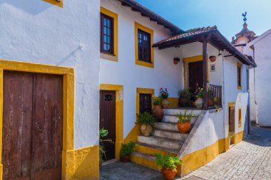 Beyaz yüzlü, sarı süslemeli ve terracotta kiremitli geleneksel bir Portekiz evi. Ev ahşap bir kapı, birden fazla pencere ve saksı bitkileriyle süslenmiş bir balkona çıkan taş bir merdiven içeriyor.