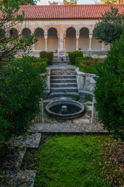Büyük yeşil yapraklı bitkiler, taş patikalar ve bahçeye çıkan taş kemer de dahil olmak üzere çeşitli bitkilerle dolu huzurlu bir manastır bahçesi.