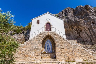 Kırmızı kapılı, küçük, bembeyaz bir şapel ve kayalık bir yamaç yüzüne kurulmuş mavi ve beyaz bir kiremit.