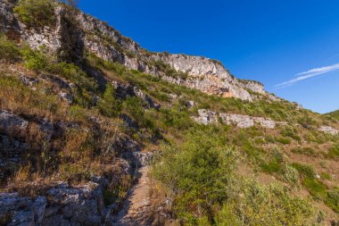 Her iki tarafında sarp kayalıklar ve yemyeşil bitki örtüsü olan engebeli bir arazide dar bir patika yol alır.