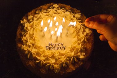 Bir el, beyaz kremayla süslenmiş bir doğum günü pastasının üzerine mum yakar ve 