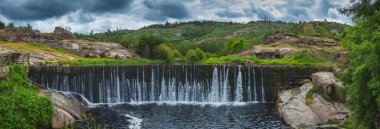 Yükselen bir şelalenin panoramik manzarası taştan bir barajın üzerinde, yemyeşil ve engebeli bir arazi tarafından çerçevelenmiş karamsar, bulutlu bir gökyüzü altında.