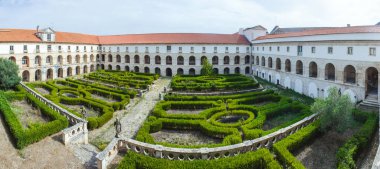 Tarihi bir manastırın içindeki manastır bahçesinde karmaşık çitler, taş geçitler ve arka planda kırmızı kiremitli çatısı olan büyük bir bina var.