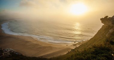 Geniş bir kumsal ve sakin okyanus dalgaları üzerinde huzurlu, altın bir gün batımı.