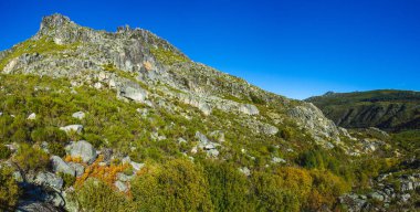 canlı mavi gökyüzünün altında engebeli, kayalık bir dağ manzarası. Arazi büyük kayalar, yeşil bitki örtüsü ve çalılarla kaplı, vahşi, doğal bir his veriyor.