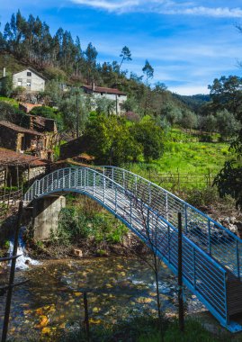 Taşra sahnesinde, her iki taraftaki yemyeşil alanları birbirine bağlayan hafif akan bir akarsu boyunca uzanan metal bir yaya köprüsü var.