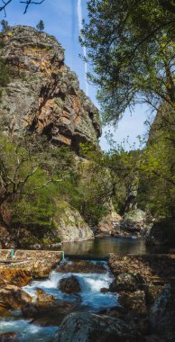 Sakin bir nehrin üzerinde yükselen engebeli bir kaya oluşumunun ön planda çağlayan küçük bir şelalesi var.