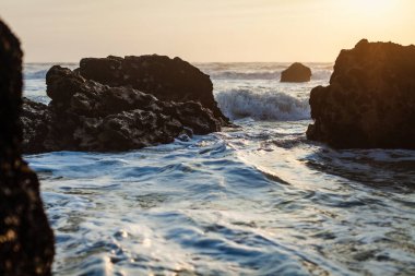 Büyük, engebeli kayalar okyanustan yükselir, dalgaların üzerine sıcak, altın bir ışık saçan batan güneşi çerçeveler.