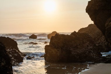 Büyük, engebeli kayalar okyanustan yükselir, dalgaların üzerine sıcak, altın bir ışık saçan batan güneşi çerçeveler.