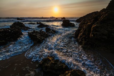 Kıyı günbatımı sahnesi, dalgalar açık denizden akın ediyor, karanlık ve engebeli kayalar arasındaki boşluğu dolduruyor..