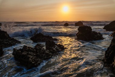 Kıyı günbatımı sahnesi, dalgalar açık denizden akın ediyor, karanlık ve engebeli kayalar arasındaki boşluğu dolduruyor..