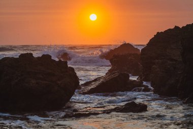 Gün batımı sahnesi kayalara çarpan bir dalganın güçlü hareketini yakalar. Güneş ufukta batarken havaya su püskürtür.