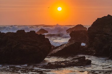 Gün batımı sahnesi kayalara çarpan bir dalganın güçlü hareketini yakalar. Güneş ufukta batarken havaya su püskürtür.