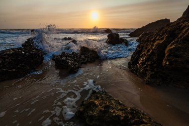 Kıyı günbatımı sahnesi, dalgalar açık denizden akın ediyor, karanlık ve engebeli kayalar arasındaki boşluğu dolduruyor..