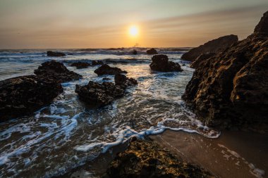 Kıyı günbatımı sahnesi, dalgalar açık denizden akın ediyor, karanlık ve engebeli kayalar arasındaki boşluğu dolduruyor..