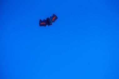 Canlı bir mavi gökyüzünde deniz kıyısında yükselen kırmızı işaretli çok kanatlı bir uçurtma var.