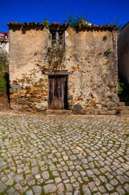 Ahşap kapılı yıpranmış bir taş bina temiz mavi gökyüzünün altında yemyeşil bir yeşil ile çevrili bir kaldırım taşı caddesinde duruyor.