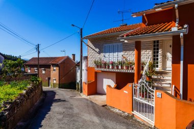 Kiremit kaplı çatılı parlak turuncu bir ev ve çiçek dolu bir balkon berrak mavi gökyüzünün altında yeşil bir cadde boyunca duruyor.