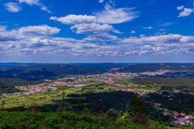 Parlak mavi bir gökyüzünün altında, dağınık yerleşim yerleri, yemyeşil ve uzak tepelerin geniş bir manzarası.