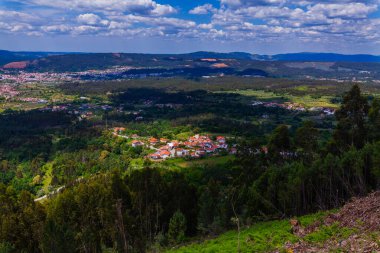 Parlak mavi bir gökyüzünün altında, dağınık yerleşim yerleri, yemyeşil ve uzak tepelerin geniş bir manzarası.