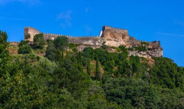 Yoğun yeşilliklerle çevrili bir tepenin üzerine tünemiş görkemli bir taş kale ve açık mavi bir gökyüzüne karşı kurulmuş.
