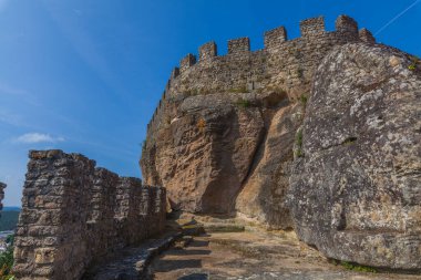 Çatlamış kuleleri ve yosun kaplı yüzeyleri olan engebeli taş bir duvar açık mavi bir gökyüzüne karşı durmakta, tarihi bir Portekiz kalesinin mimarisini gözler önüne sermektedir.