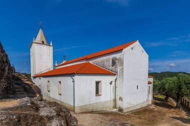 Kırmızı kiremitli ve çan kulesi olan küçük beyaz bir kilise kayalık arazi ve yeşilliklerle çevrili açık mavi bir gökyüzüne karşı duruyor.