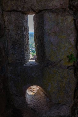 Kale duvarındaki dar bir boşluk, yosun kaplı duvarlarla çerçevelenmiş ve uzak yeşillik manzarasına yol açıyor.