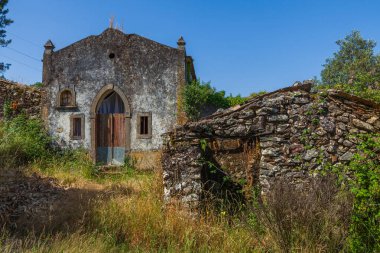Kırsal bir kemer yolu ve ahşap kapılı eski bir taş bina, açık mavi gökyüzünün altındaki taş bir duvarla çevrelenmiş, fazla gelişmiş bitki örtüsünün arasında duruyor..