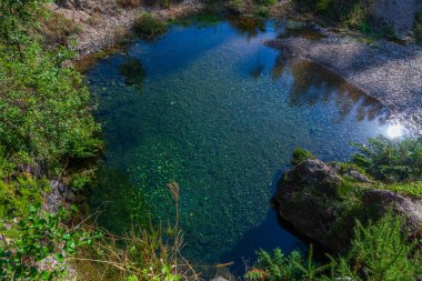 Kristal berraklığında suyu olan sakin bir doğal havuz, engebeli bir kaya ve bitki örtüsünün içine yerleştirilmiş yeşil ve güneş ışığını yansıtır.