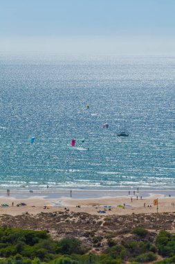 Güneşli bir günde kıyı şeridi tarafından çerçevelenmiş, açık mavi okyanusta uçurtma sörfçüleriyle dolu hareketli bir kumsal manzarası.