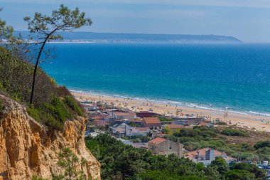 Geniş, yüksek açılı manzaralı yeşil bir vadi ve kıyı kasabasına bakan geniş kumlu bir sahil, berrak bir gökyüzünün altında sakin bir mavi okyanusla buluşuyor.