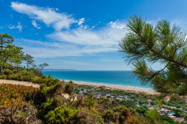 Sahil kasabasına bakan canlı, yüksek açılı bir manzara ve parçalı bulutlu bir gökyüzünün altında sakin mavi bir okyanusa açılan geniş kumlu bir sahil.