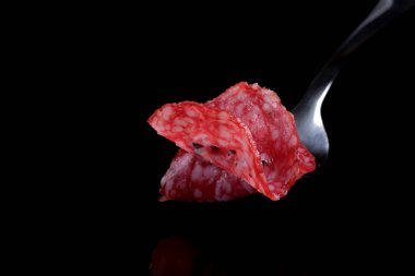 Thinly sliced delicious fragrant salami sausage on a steel fork close-up on a black mirror background