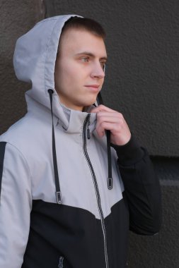 Kapüşonlu kısa saçlı, siyah taştan bir duvara dayalı genç bir adamın yakın plan portresi.