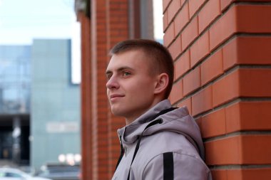 Kısa saçlı, açık renk ceketli, kırmızı tuğlalı duvarın yanında kapüşonlu genç bir adamın portresi.