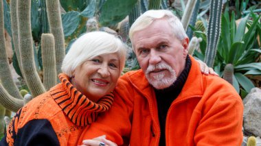 Turuncu kazaklarla botanik bahçesindeki yeşil bitkilerin arka planına karşı aşık yaşlı, gri saçlı iyimser bir çift.