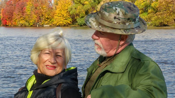高齢者の白髪のカップル 軍事化されたカーキの服の男 黒いウインドブレーカーの女性 秋には川の堤防の街での肖像 — ストック写真