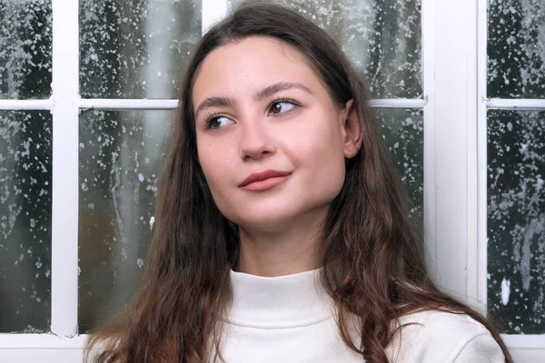冬の窓を背景に白いセーターを着た美しいかわいい若い女性の肖像画 — ストック写真