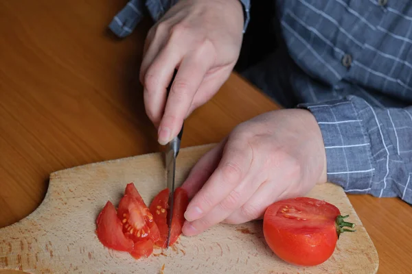 Slicing a red ripe tomato. An elderly woman\'s hands hold a tomato for slicing on a table in the kitchen. Close-up plan.