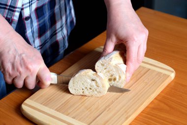 Yaşlı bir kadının elleri, kahverengi bir masanın mutfağında yüzsüz bir bıçakla beyaz buğday ekmeği kesiyor.
