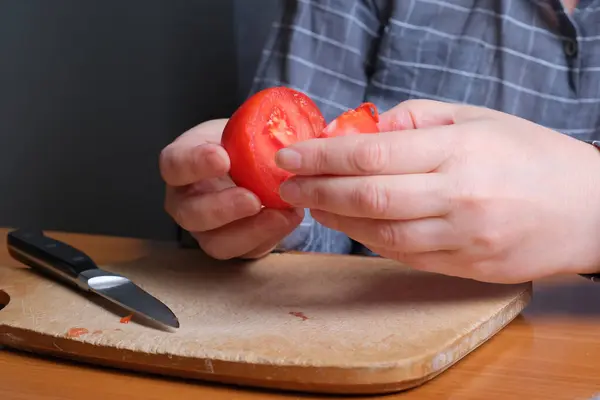 Hands of an elderly woman cutting a red ripe tomato on a cutting board for a snack in the kitchen at a brown table, no face, close-up