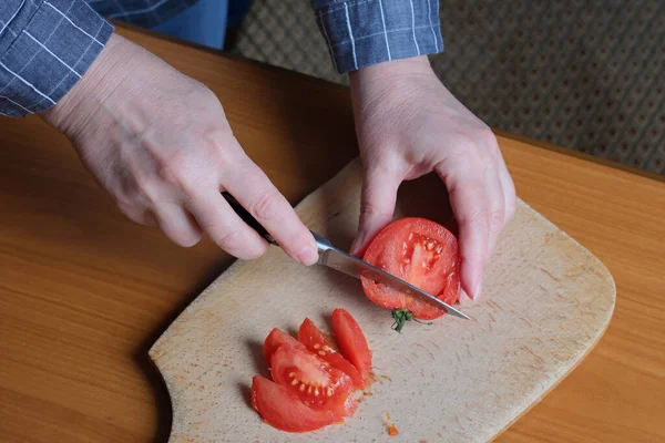 Hands of an elderly woman cutting a red ripe tomato on a cutting board for a snack in the kitchen at a brown table, no face, close-up