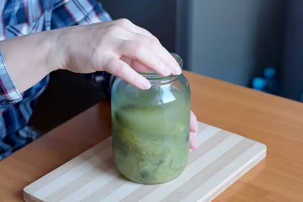 一位老妇人的手从一个罐子里拿出美味的自制黄瓜 在一个没有脸的棕色桌子旁边的厨房里当作小吃来切碎 — 图库照片
