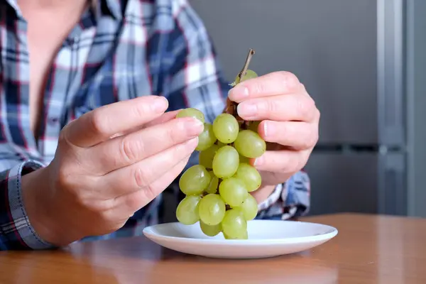 The hands of an elderly woman holds a small brush of white seedless grapes and displays them in the kitchen at a brown table, without a face, close-up